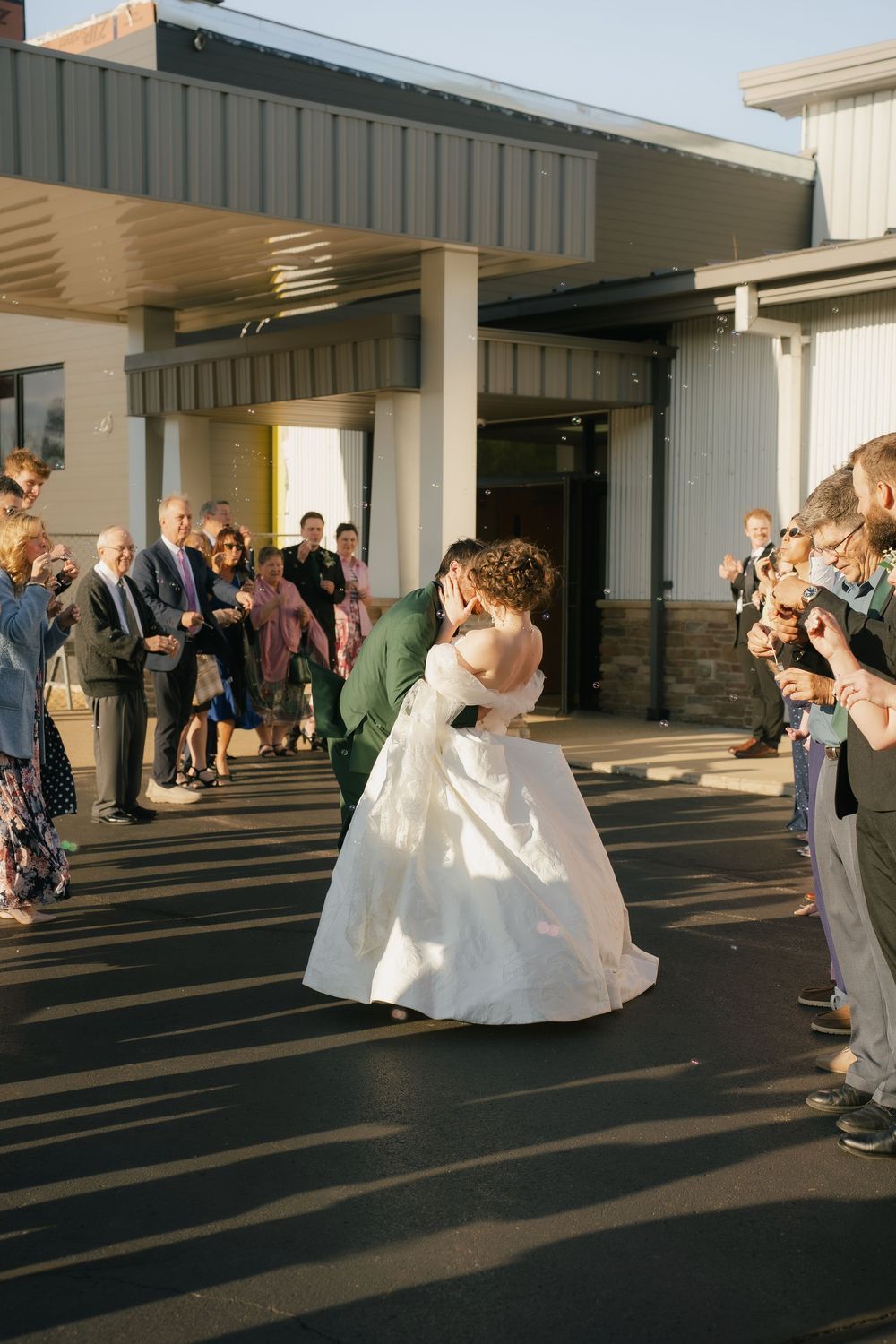 Bride and groom share a kiss outside wedding venue while guests celebrate and photograph the newlyweds.