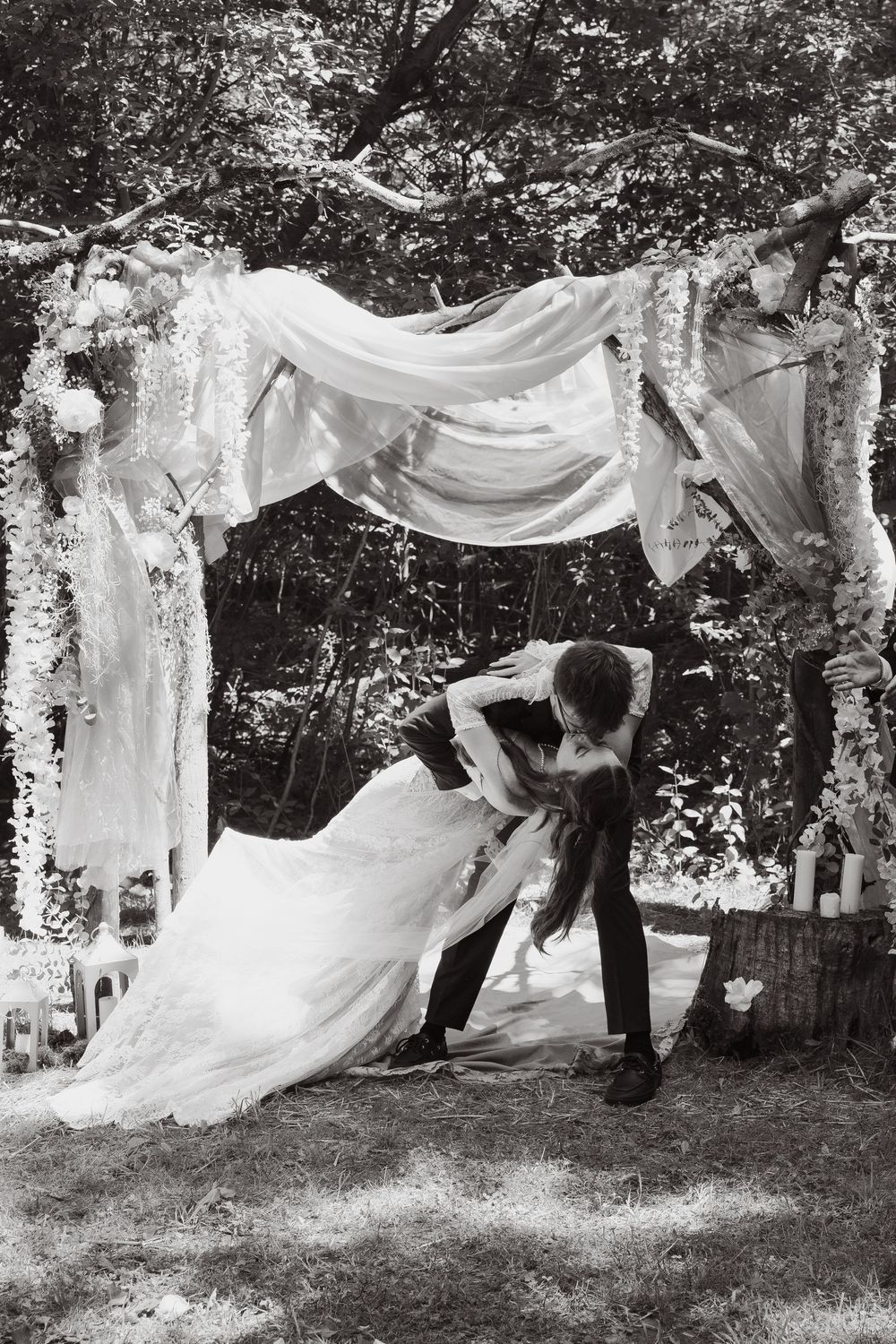 Black and white photo of bride and groom sharing a romantic dip kiss under a rustic floral arch outdoors.