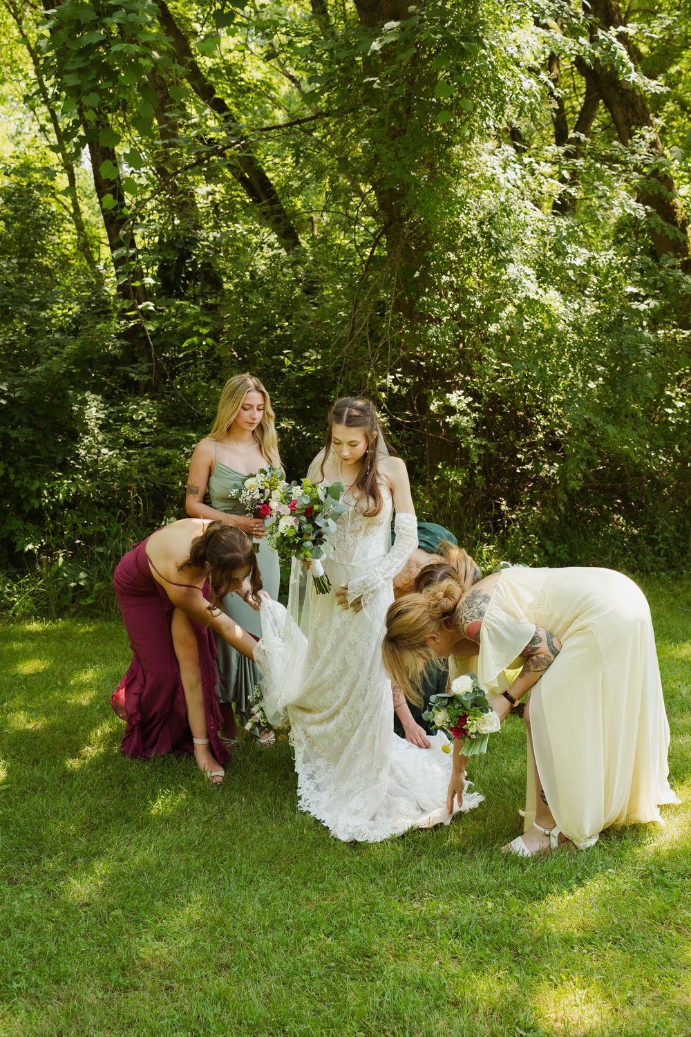 Bridesmaids in colorful gowns help bride with white lace dress on green lawn surrounded by lush trees.