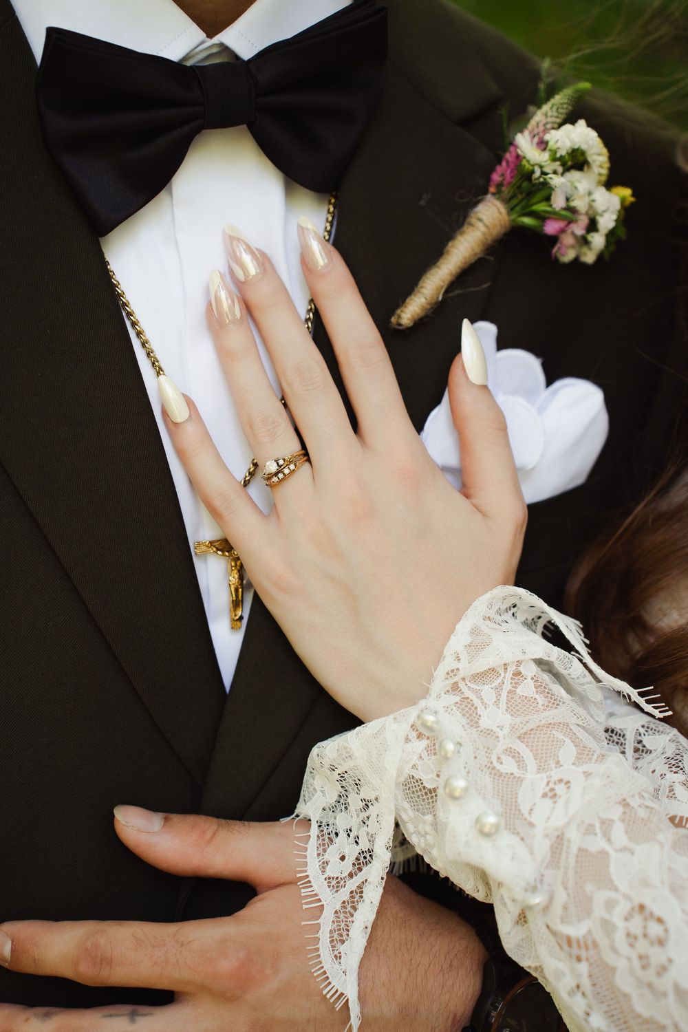 Bride's hand with gold claddagh ring and nude nails rests on groom's black tuxedo with boutonniere and bow tie.