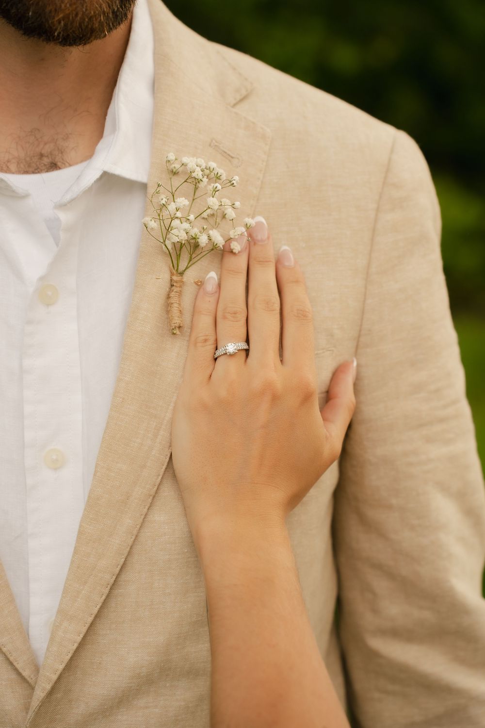 Bride's hand with diamond wedding ring resting on groom's beige linen suit jacket adorned with baby's breath boutonniere.