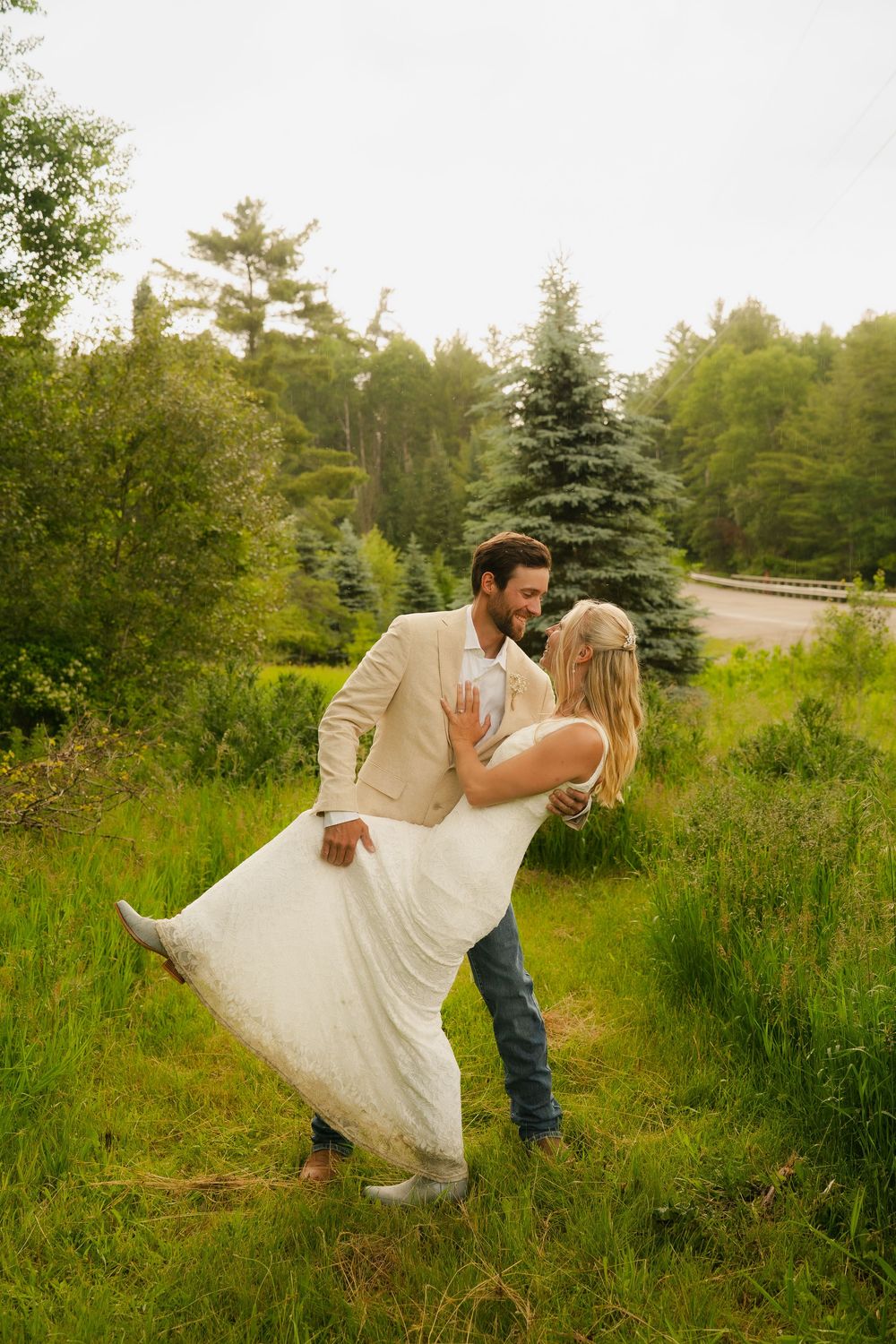 Groom in tan jacket dips bride in white dress outdoors in lush green field with evergreen trees.