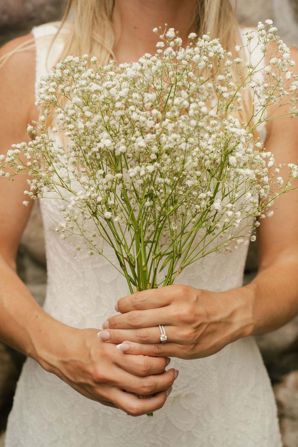 Bride in white lace dress holding baby's breath bouquet with diamond engagement ring visible on her hand.