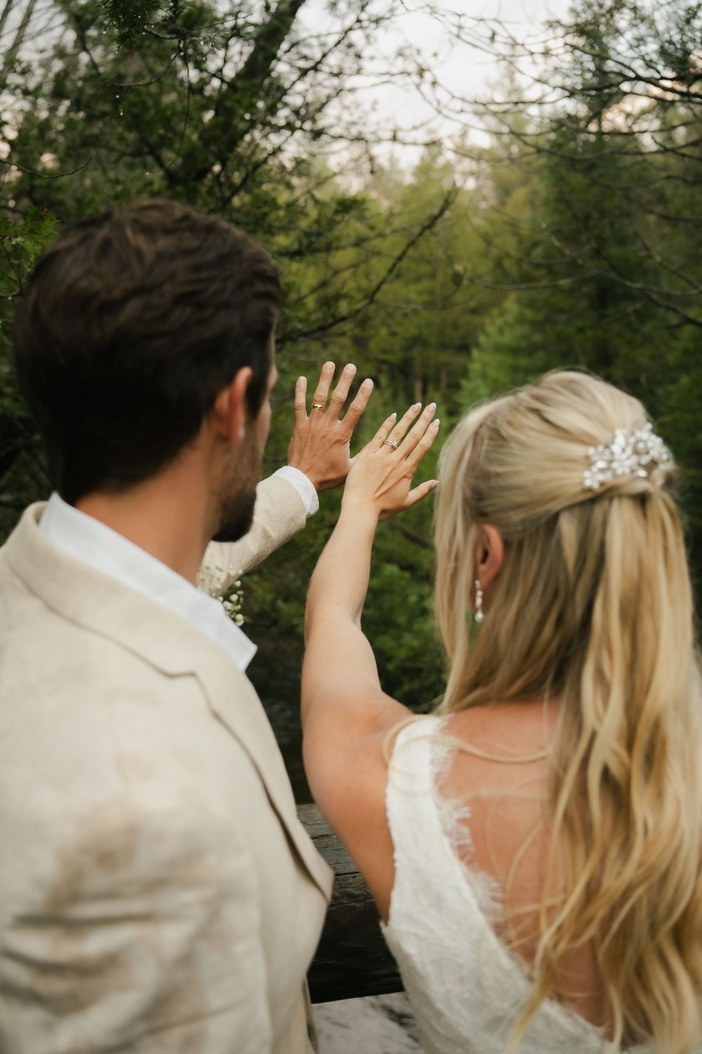 Bride and groom raising hands showing wedding rings with lush green forest backdrop on their wedding day.