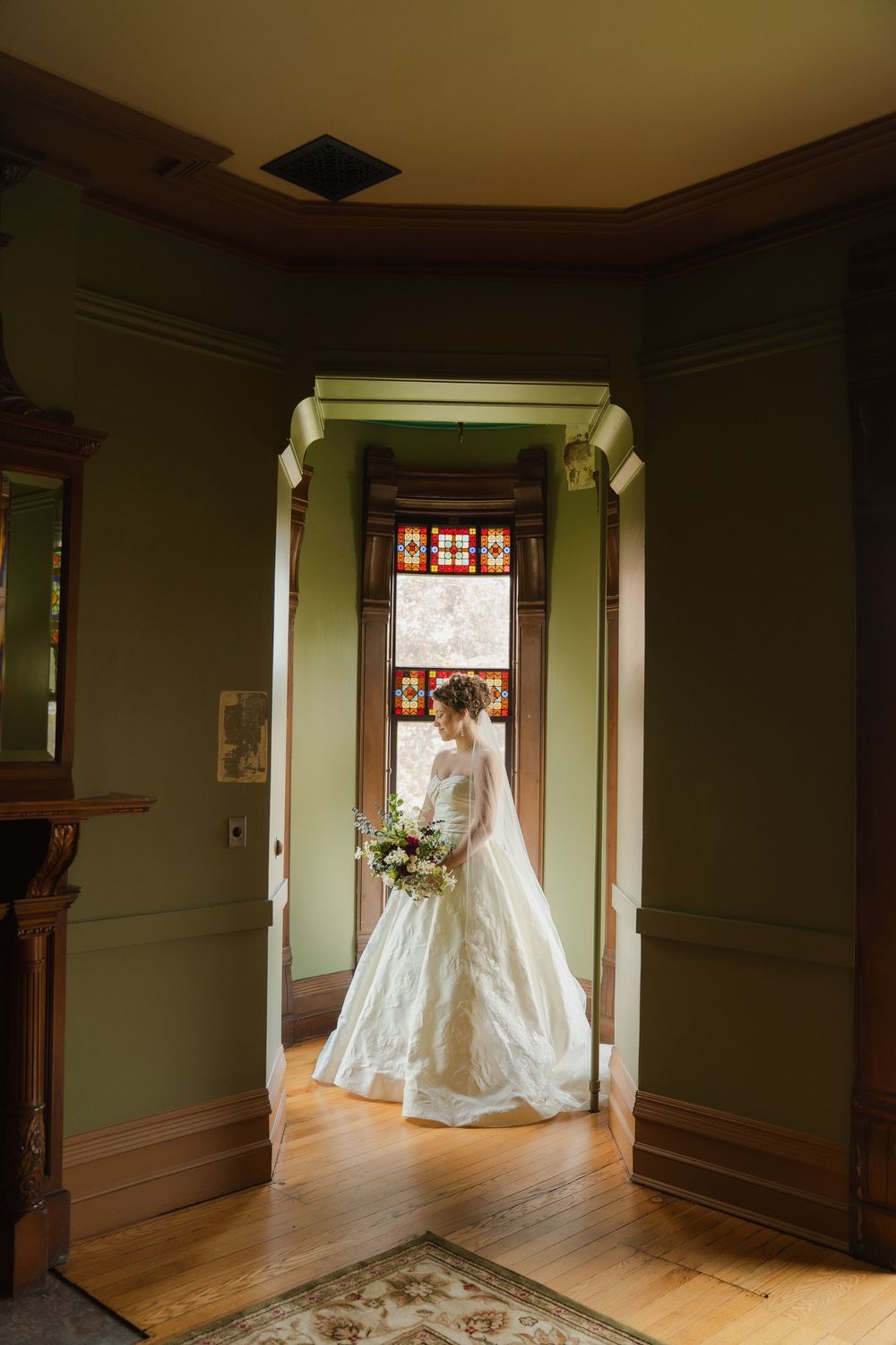 Bride in a flowing white gown holding a bouquet stands in a doorway with stained glass windows in a Victorian home.