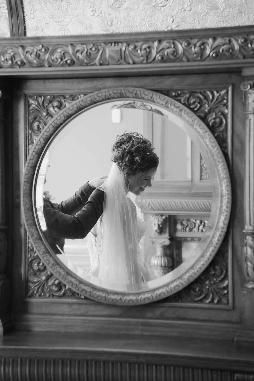Black and white photo of a bride in a wedding veil reflected in an ornate vintage round mirror.