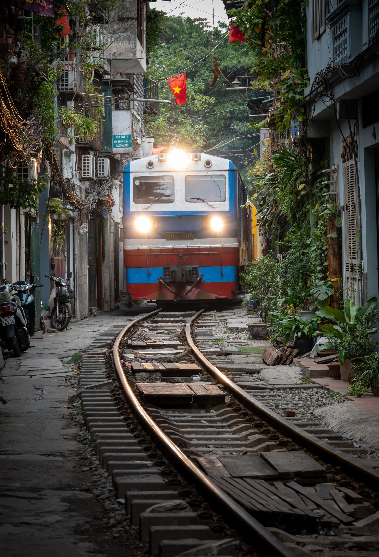 A blue train approaches through a narrow urban alley with overhanging plants and Vietnamese flags.
