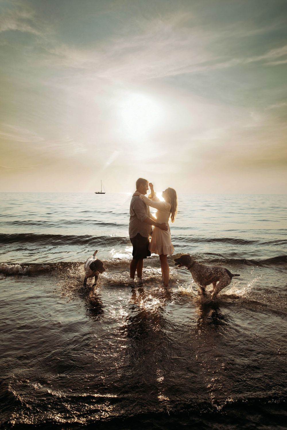 Romantic couple silhouetted against sunset ocean waves with two playful dogs splashing.