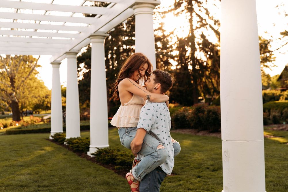 Couple sharing a romantic moment under white pergola columns in a sunlit garden, man lifting woman.