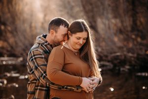 Pregnant couple embracing tenderly outdoors by a creek in warm autumn sunlight.