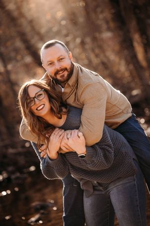Bearded man hugging smiling woman from behind in a playful piggyback pose outdoors.