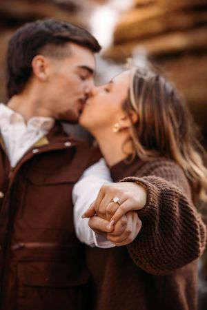 Engaged couple kissing while displaying diamond engagement ring outdoors.