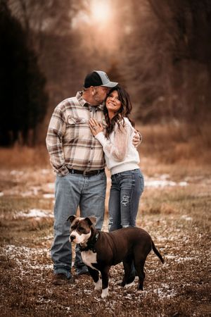 Couple embracing outdoors in winter with their brown and white pit bull dog in a snowy field.