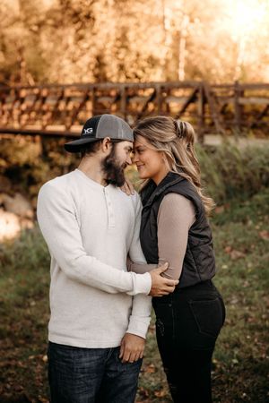 Couple sharing a tender nose-to-nose moment outdoors near a rustic bridge in autumn sunlight.