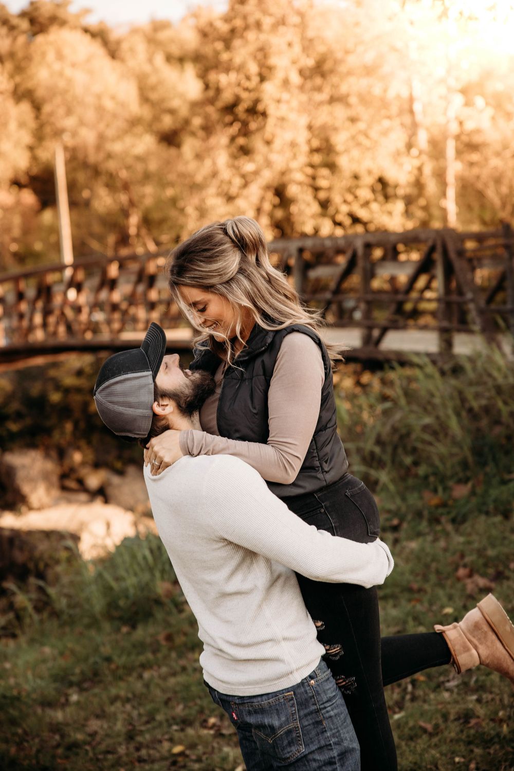Man lifting woman in a joyful embrace near a wooden bridge surrounded by fall foliage.