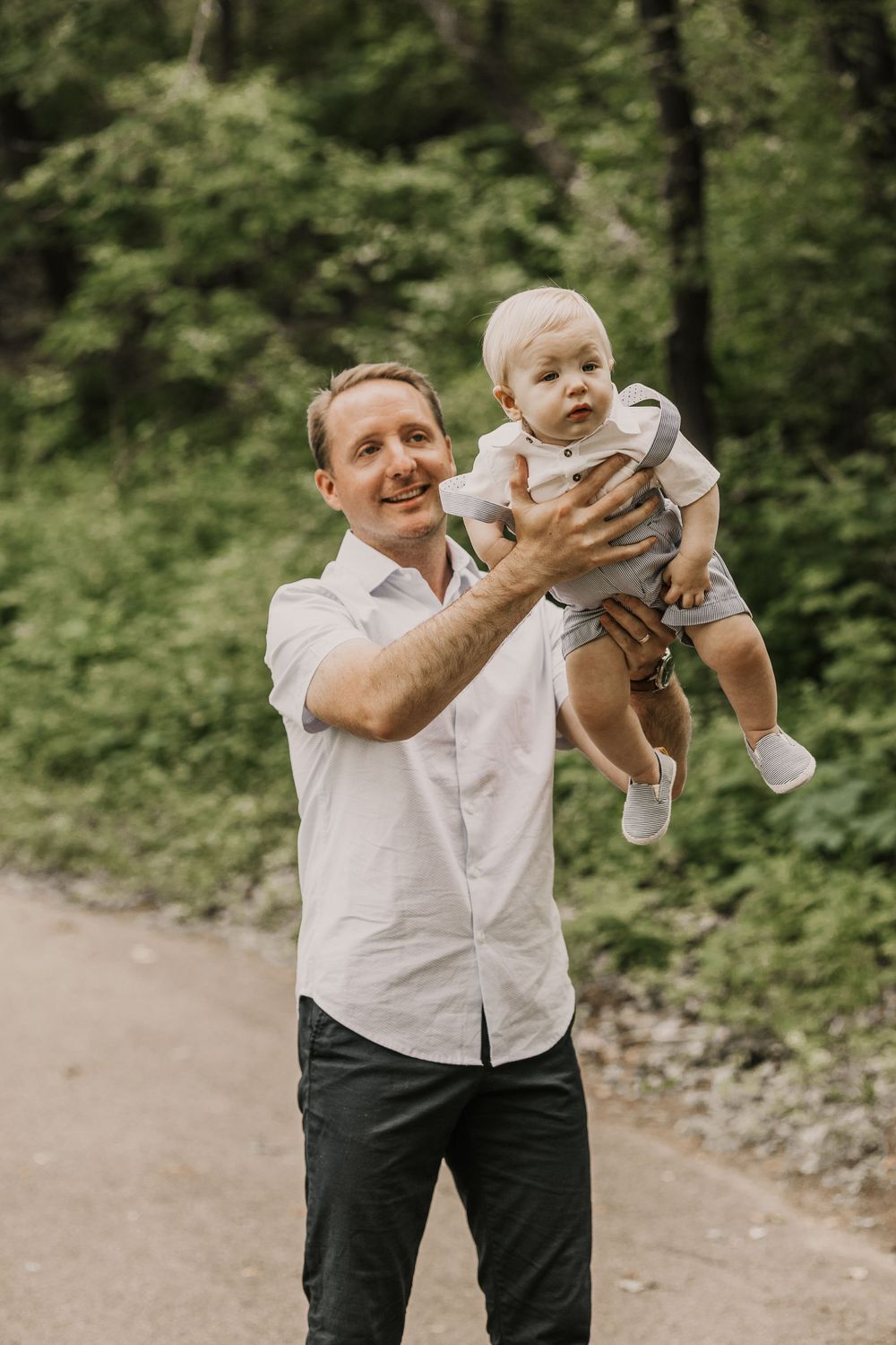 A person in a white shirt holds up a baby outdoors while smiling.