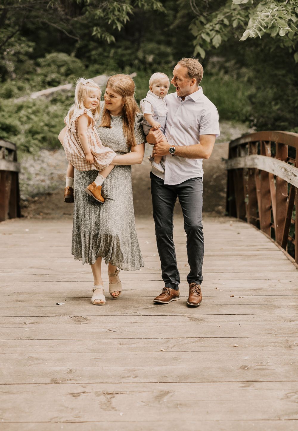 A family of four stands together on a wooden bridge in a park setting.