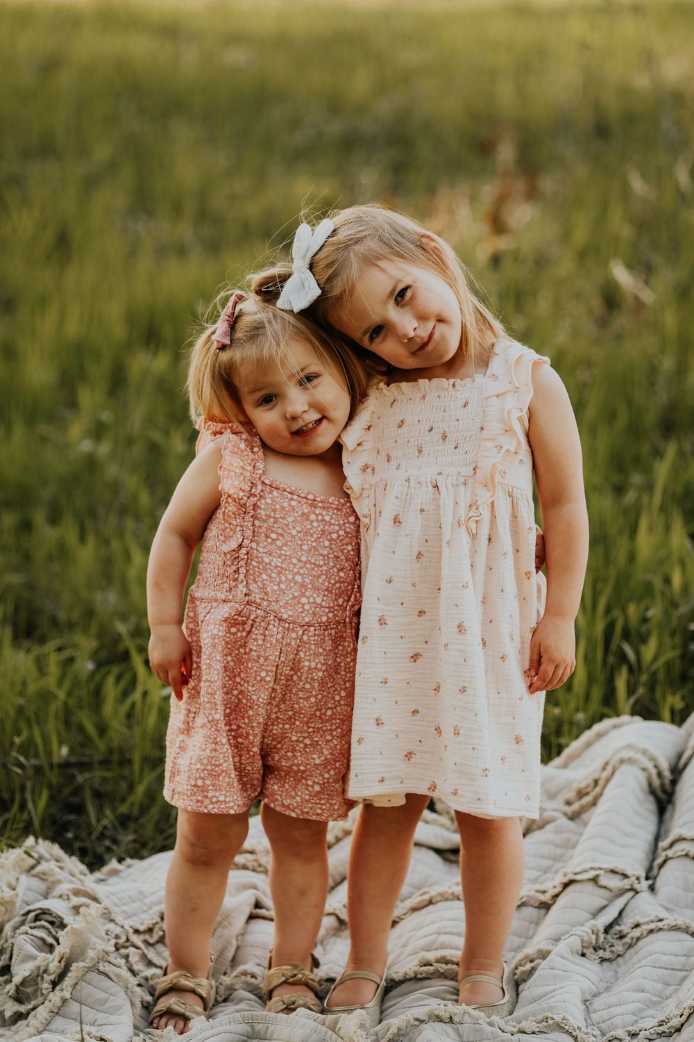Two young children in summer dresses standing together on a blanket in a grassy field at sunset.