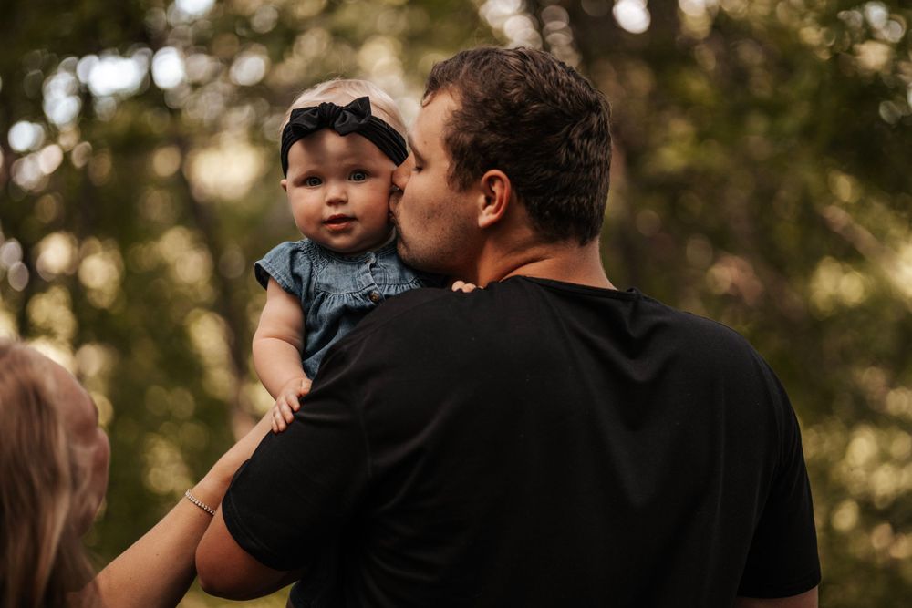 A person holds a baby wearing a navy outfit against a backdrop of trees at sunset.