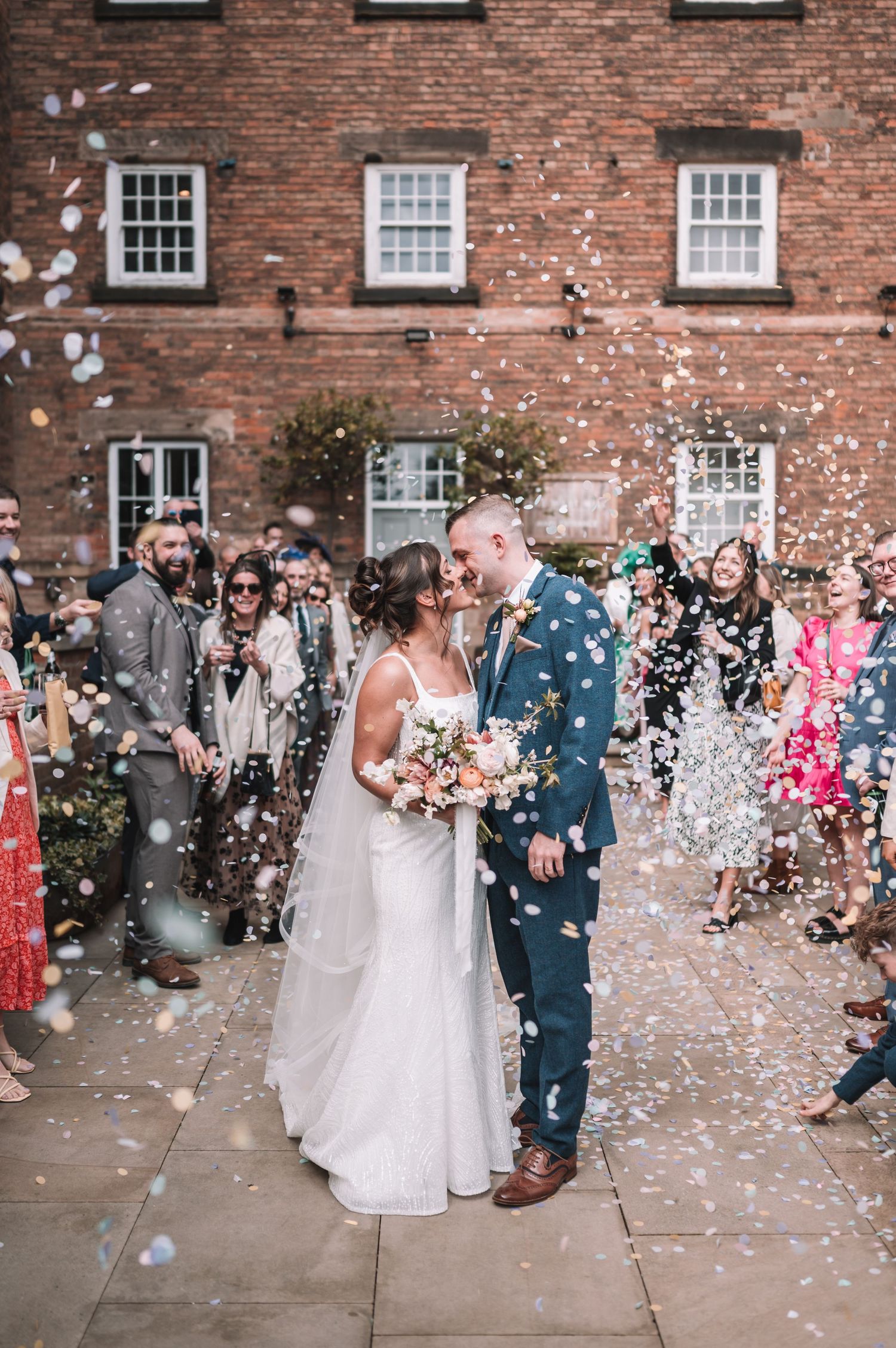 A bride and groom embrace while white confetti falls around them in front of a rustic brick building at the West Mill, Derby.