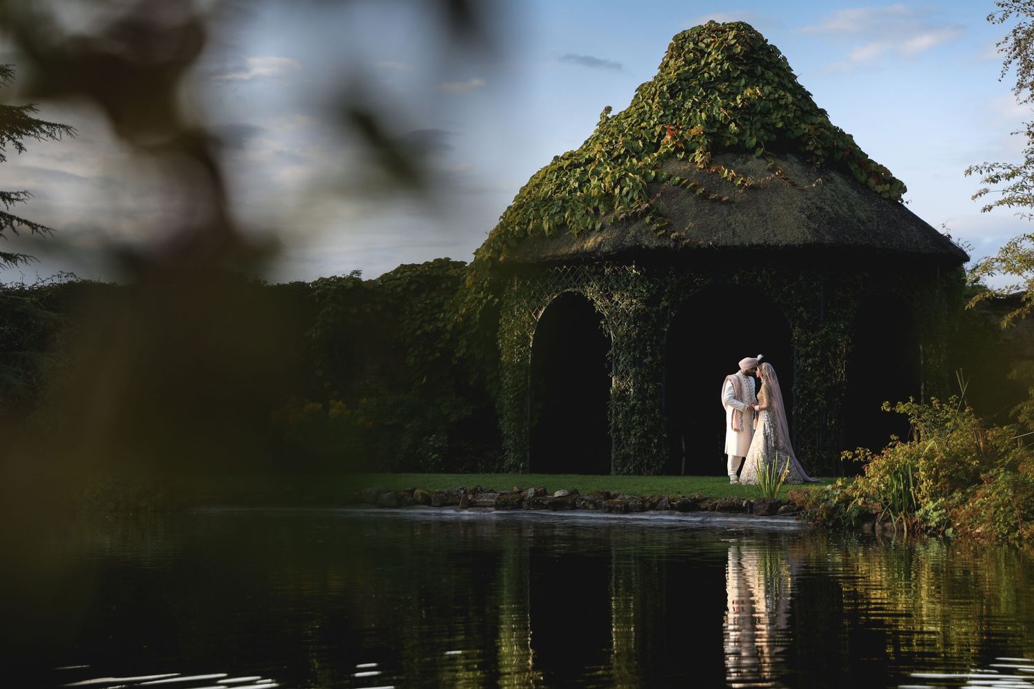 A moss-covered stone gazebo reflects in a serene pond as a couple celebrates their wedding day in an elegant pose.