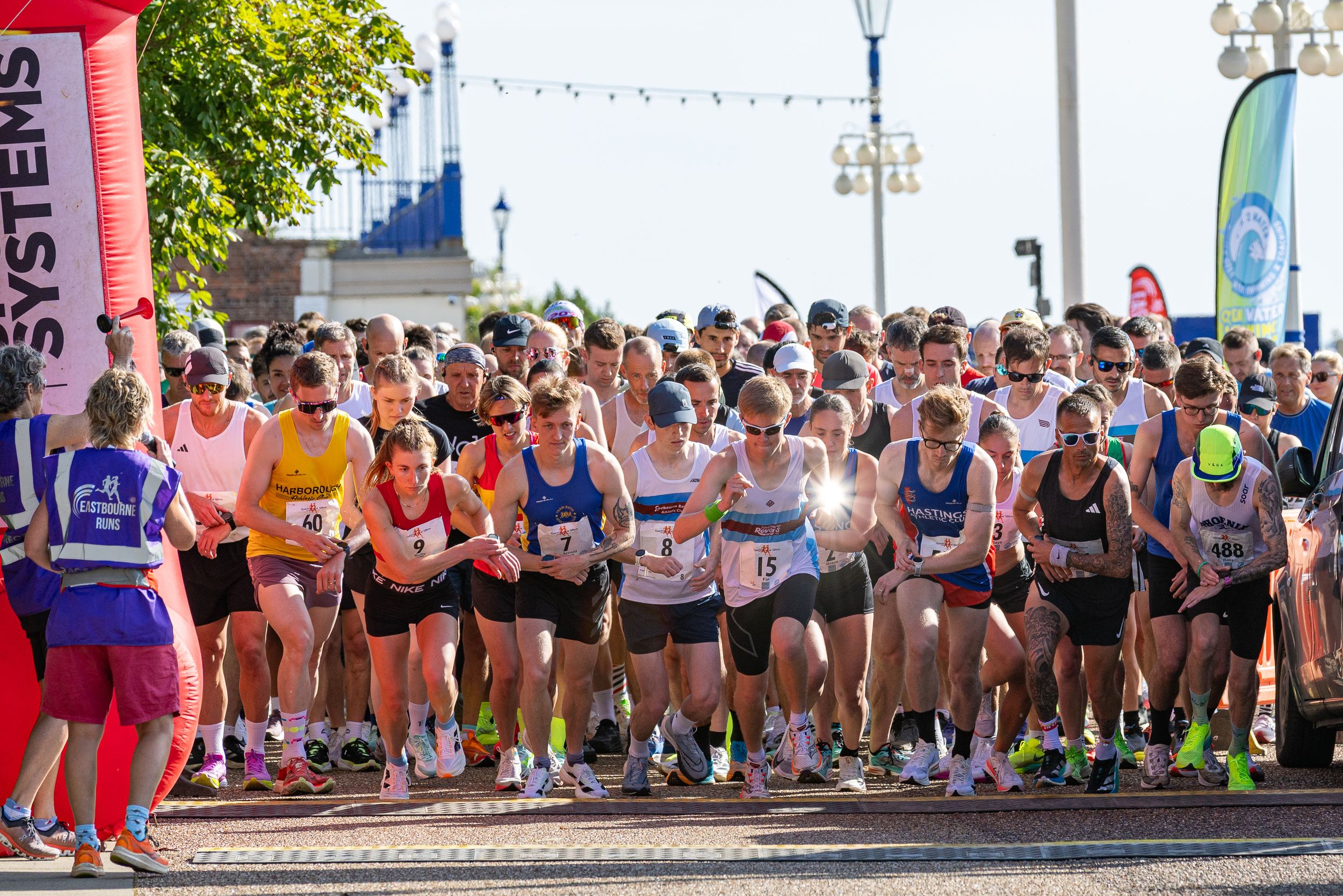 Marathon runners line up at the starting line of a summer race on a sunny day with spectators watching.