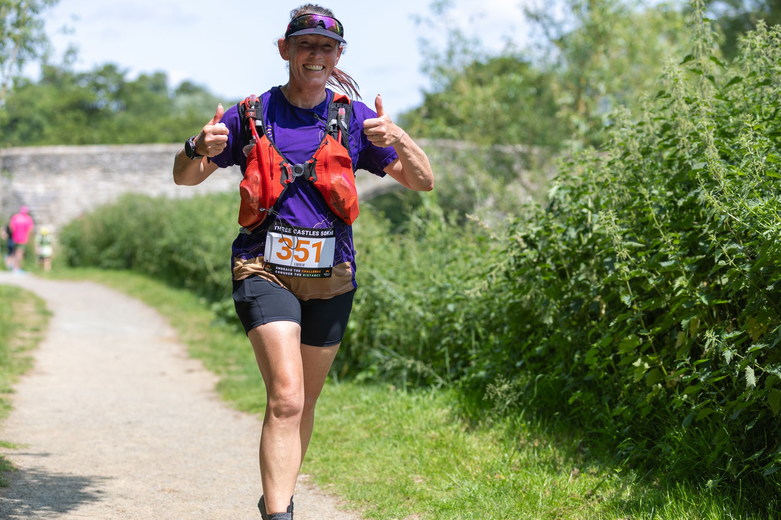 Trail runner in colorful gear gives thumbs up while jogging on sunny dirt path bordered by green vegetation.