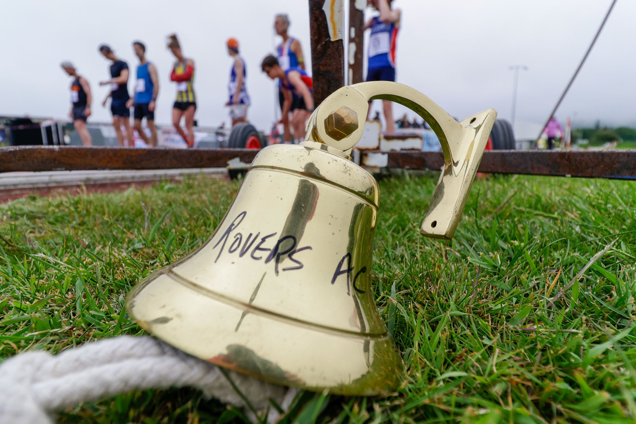 A vintage brass bell rests on grass with runners visible in the background.