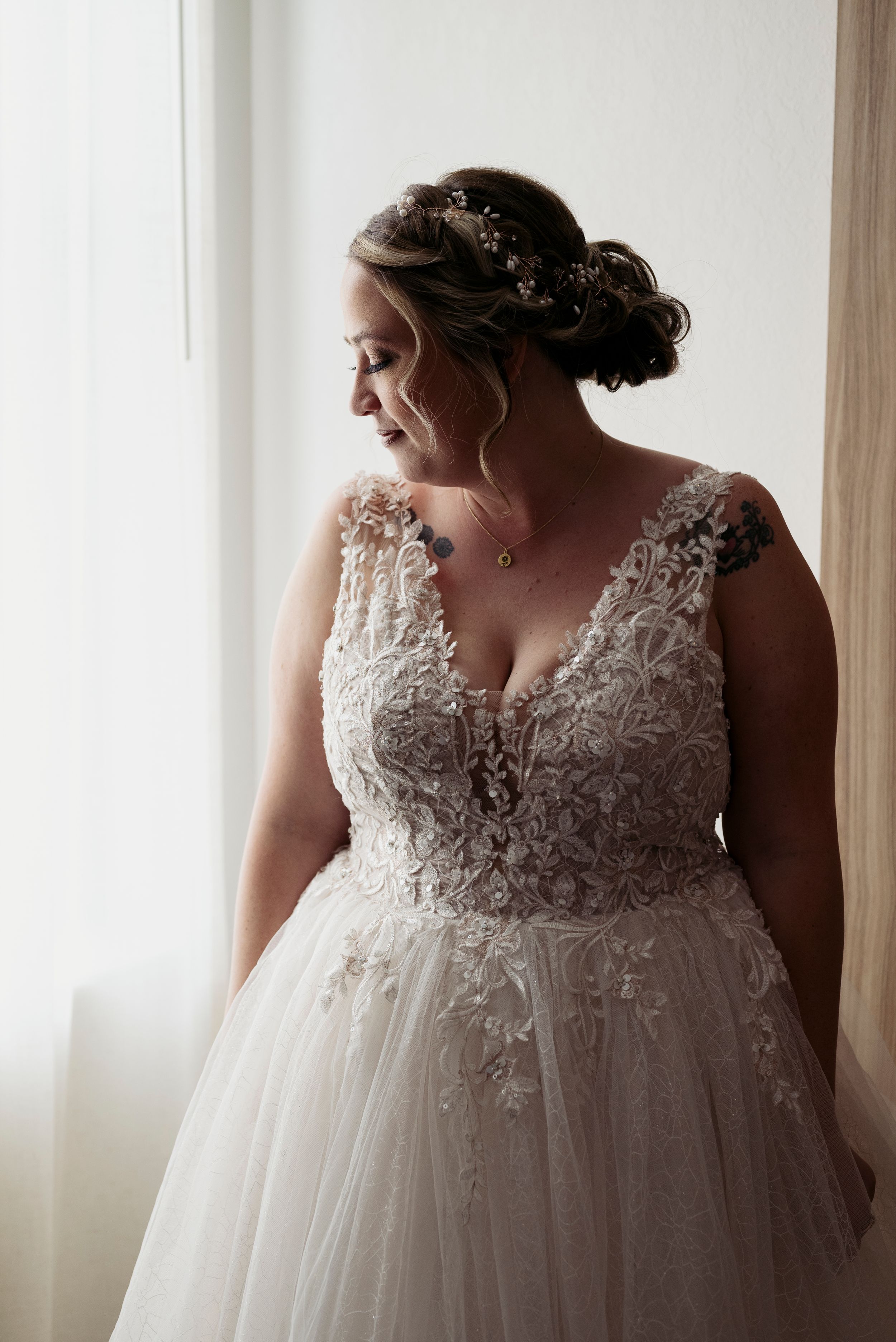 Bride in lace bodice tulle ball gown wedding dress with floral hair accessory posing by window.