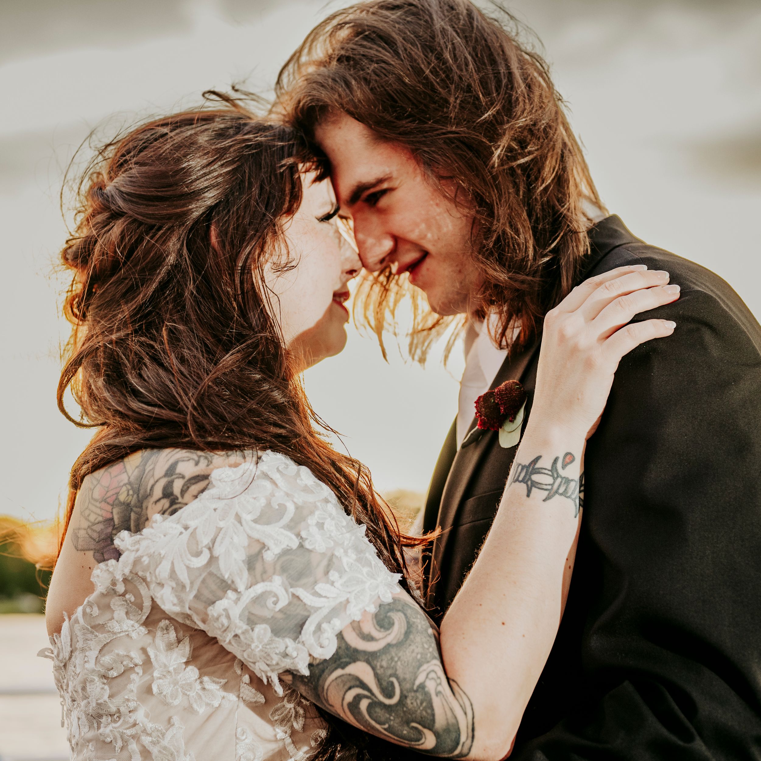 Tattooed bride and groom with long hair touching foreheads and smiling intimately at outdoor wedding.