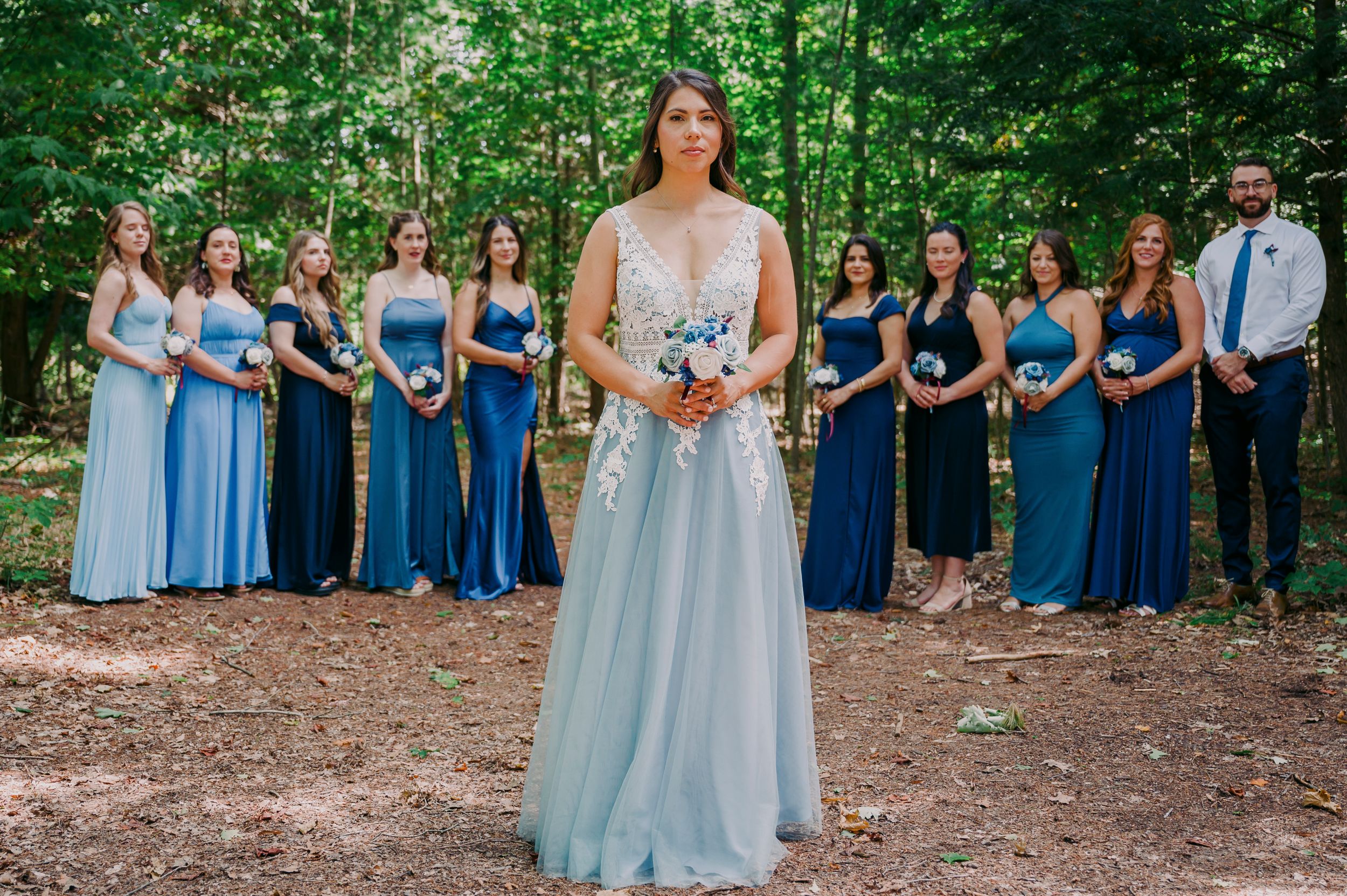 Bride in light blue lace wedding gown poses before wedding party in blue bridesmaid dresses outdoors.