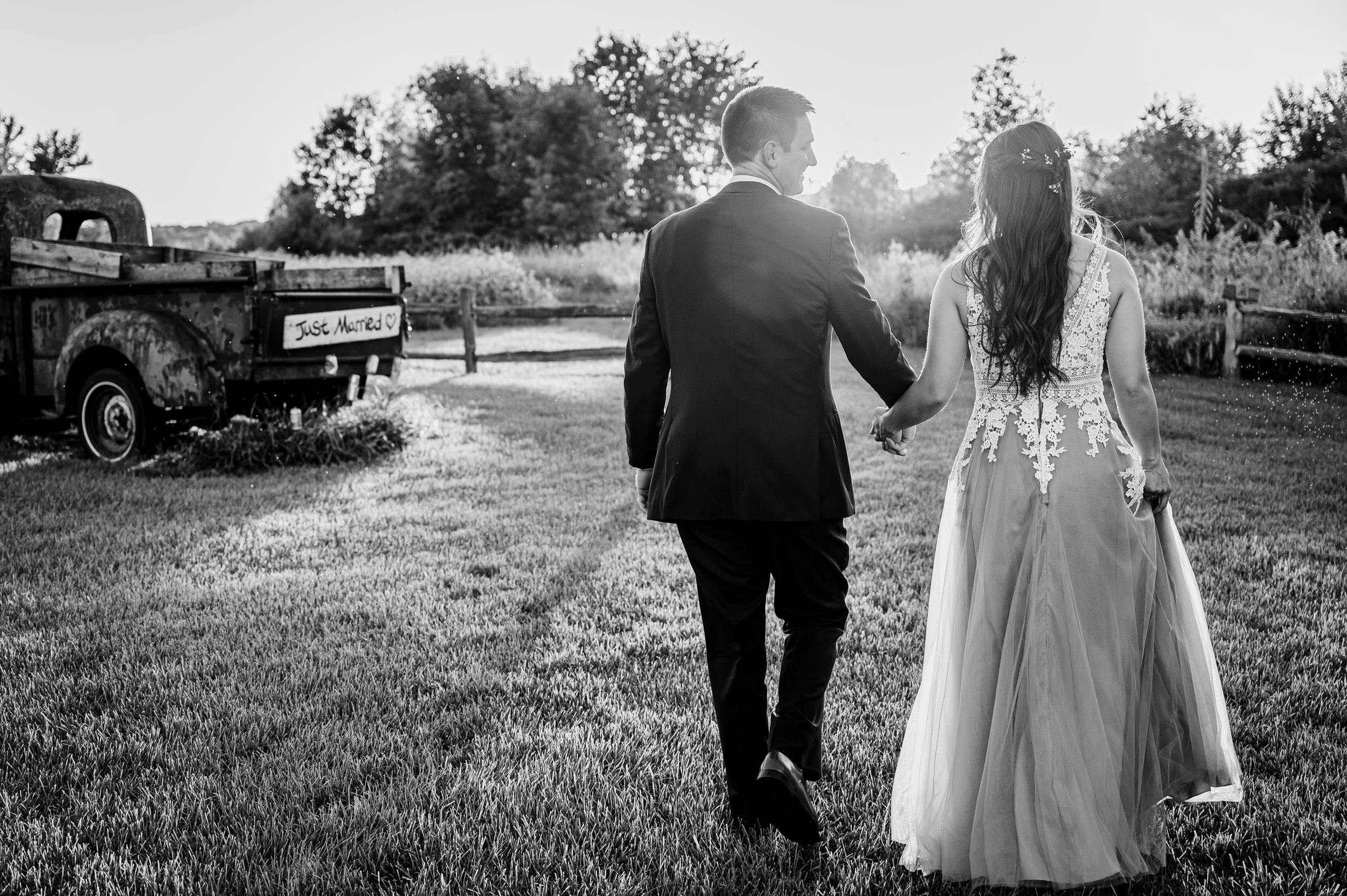 Bride and groom walking hand in hand toward vintage truck at rustic outdoor wedding in black and white.