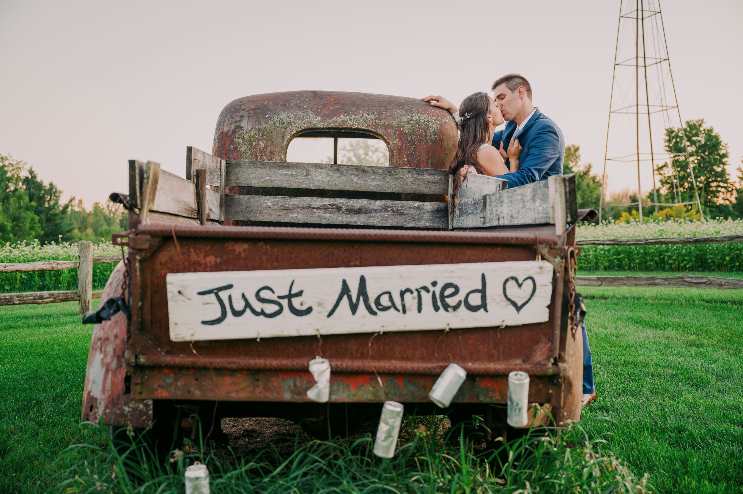 Newlyweds kissing behind rustic truck with Just Married sign at outdoor farm wedding at sunset.
