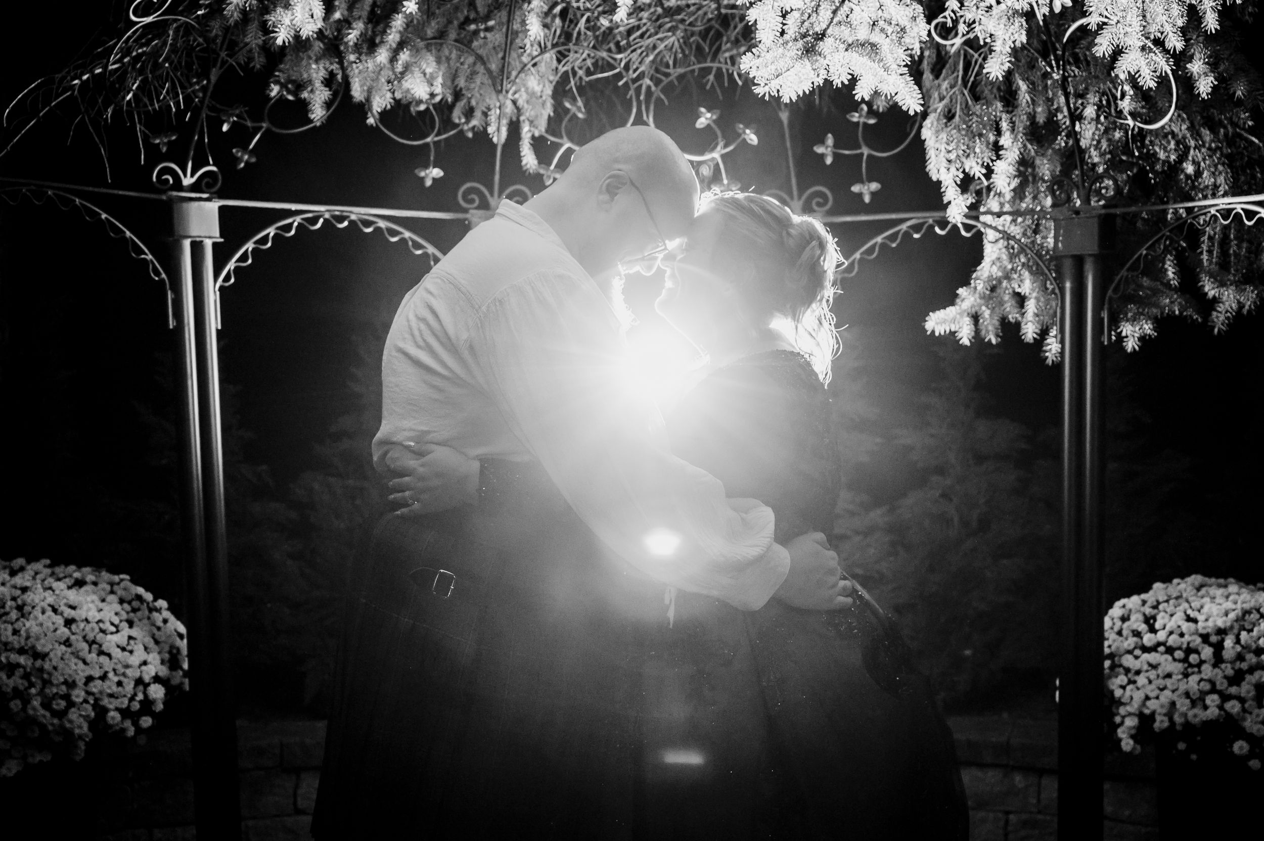 Black and white romantic photo of couple kissing under floral arch with dramatic light burst.