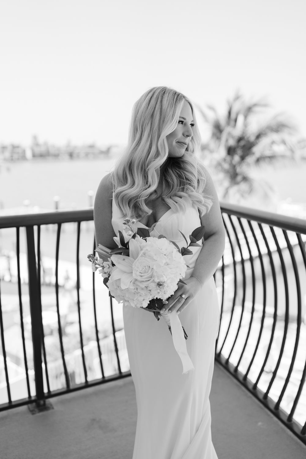Black and white photo of elegant bride in fitted sleeveless gown holding white floral bouquet on balcony overlooking palm trees.