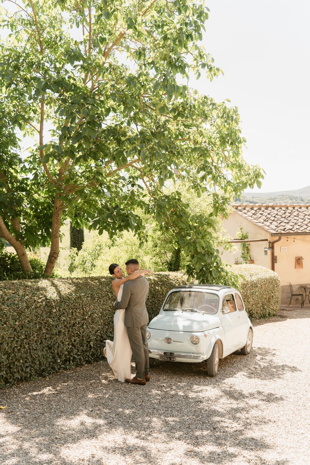 A couple shares a romantic moment next to a vintage car on a sunny day by a stone wall.