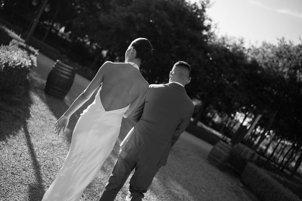 A couple in wedding attire walks together on a path while photographed from behind in black and white.