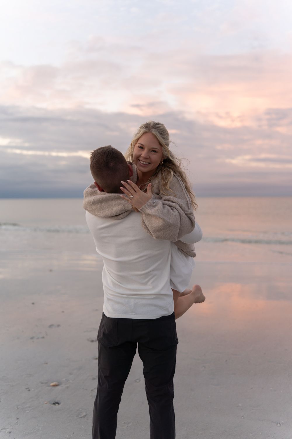 A couple embraces joyfully on a beach at sunset with gentle waves and pink clouds in the background.