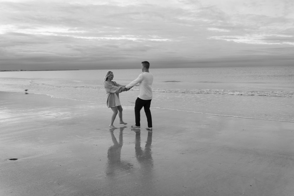 Two figures hold hands and dance together on a misty beach at sunset, creating reflections on the wet sand.