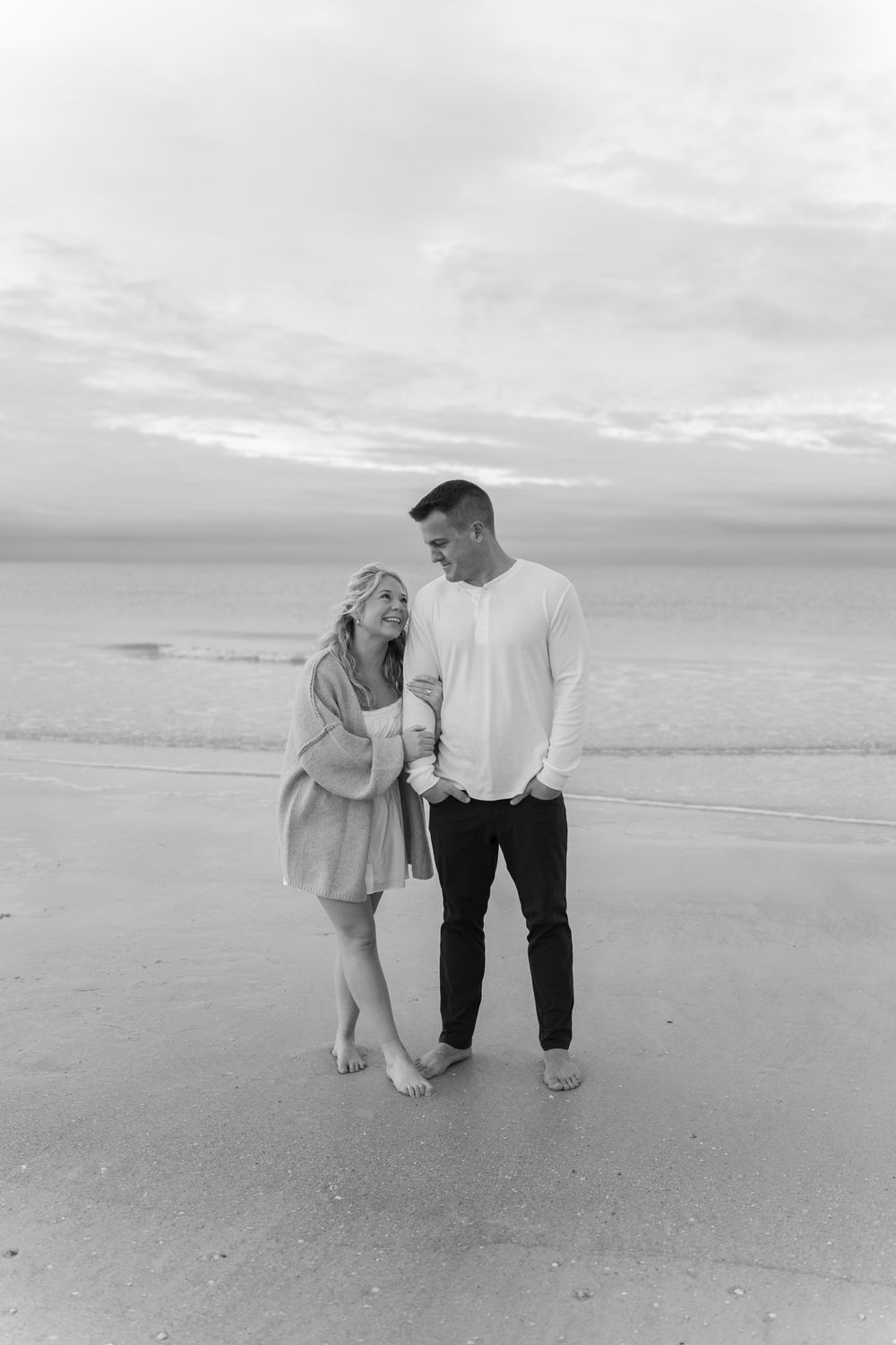 A couple shares an intimate moment while standing barefoot on a misty beach at dusk in black and white photography.