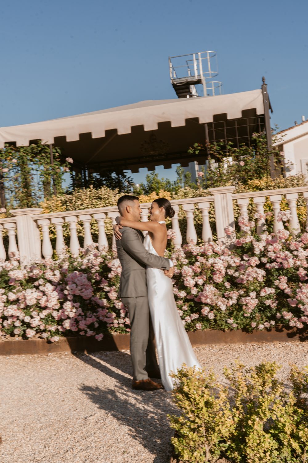 A couple embraces on a garden terrace surrounded by pink roses and elegant stone balustrades under a sunny blue sky.