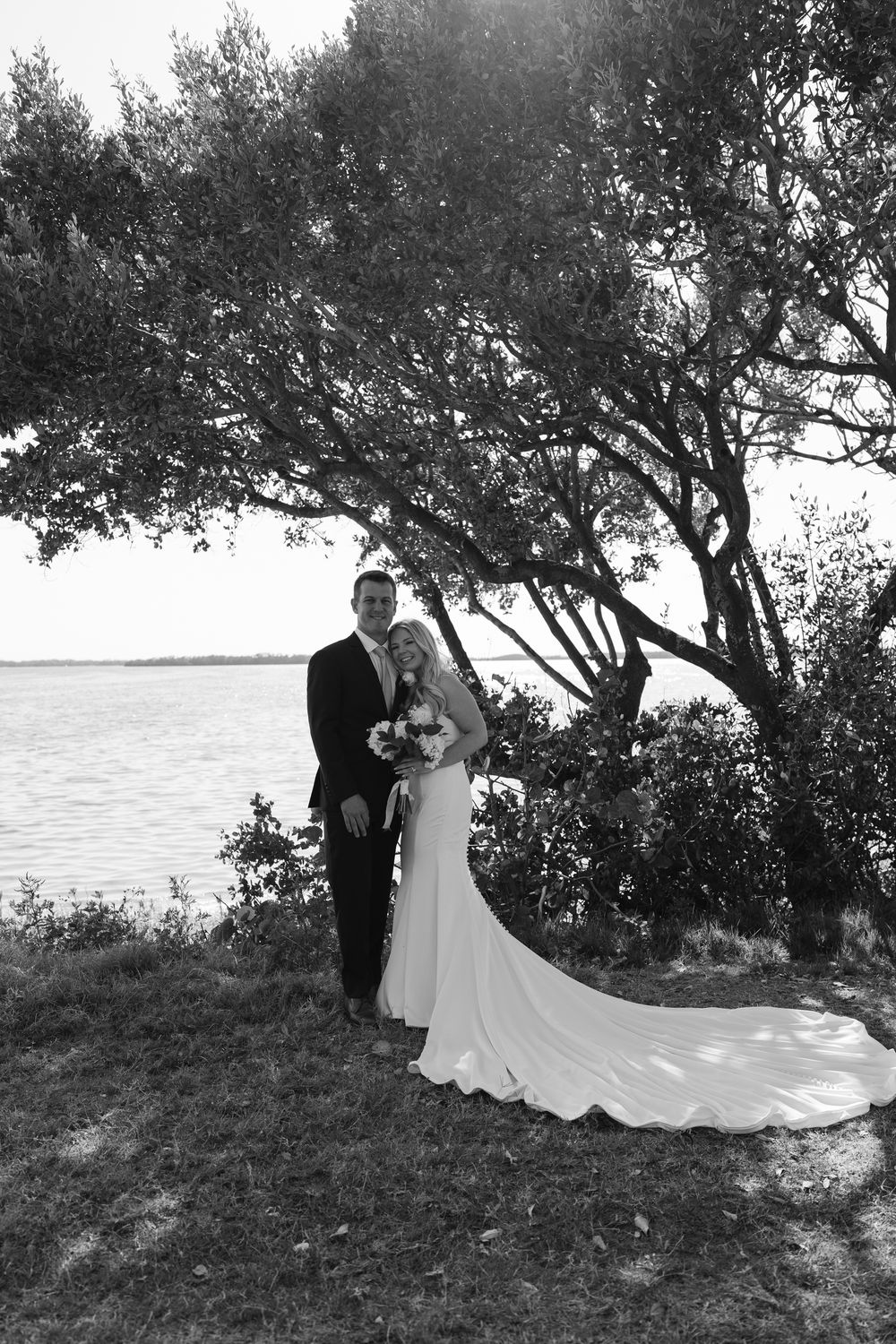 A wedding couple poses under a tree by the waterfront in an elegant black and white photograph.