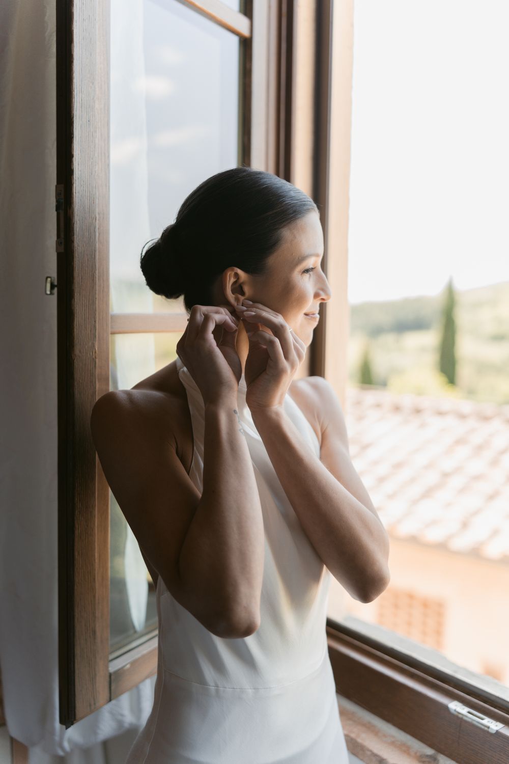 A figure in a white dress gazes thoughtfully through a sunlit window with their hands near their face.