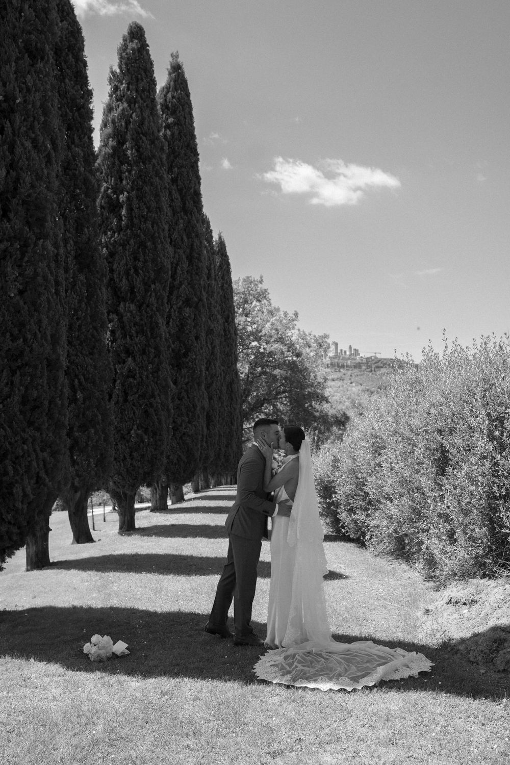 A romantic couple shares a moment on a path lined with tall cypress trees in a black and white countryside setting.