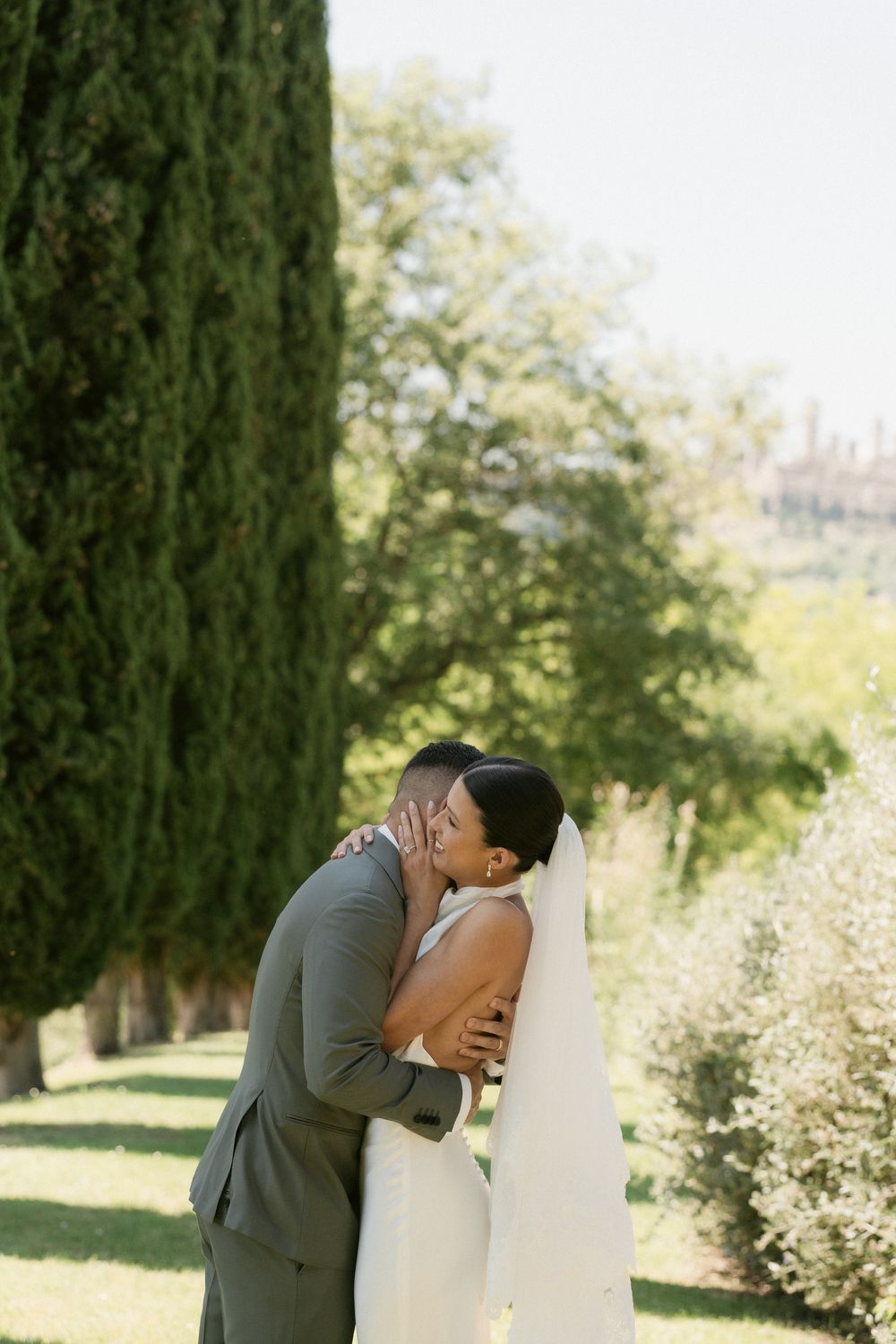 A couple shares a romantic kiss on their wedding day in a lush garden setting with cypress trees in the background.