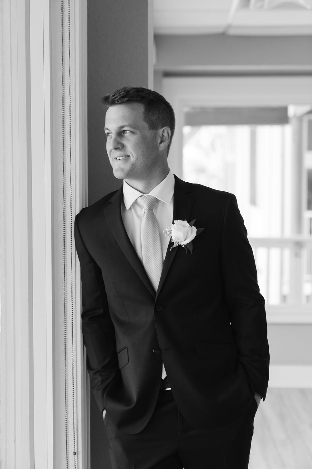 A groom in a classic black tuxedo with white boutonniere stands by a window in a black and white photograph.