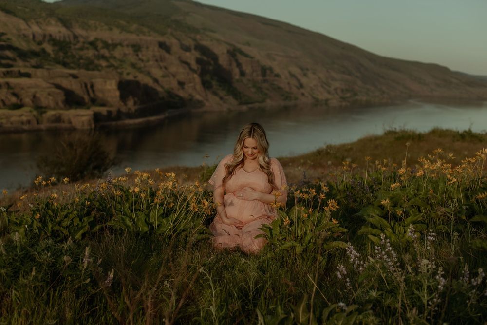 A dreamy sunset portrait in a pink dress among wildflowers overlooking a serene river valley landscape.