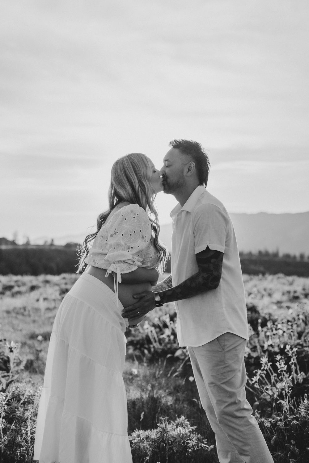A couple shares a romantic moment in a flower field during sunset, captured in black and white photography.