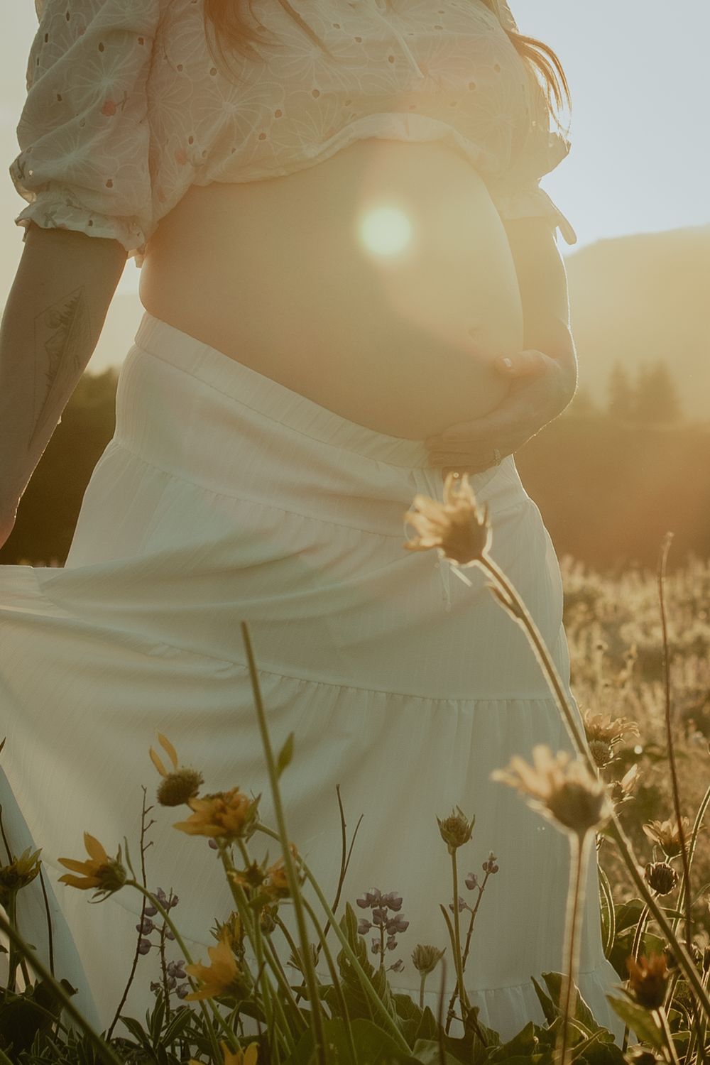 A white maternity dress silhouetted against golden sunlight with wildflowers in the foreground during sunset.