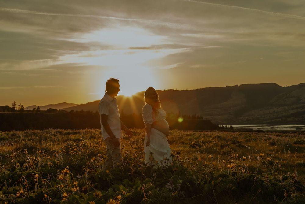 Two figures stand in silhouette against a golden sunset sky in a wildflower meadow overlooking mountains.